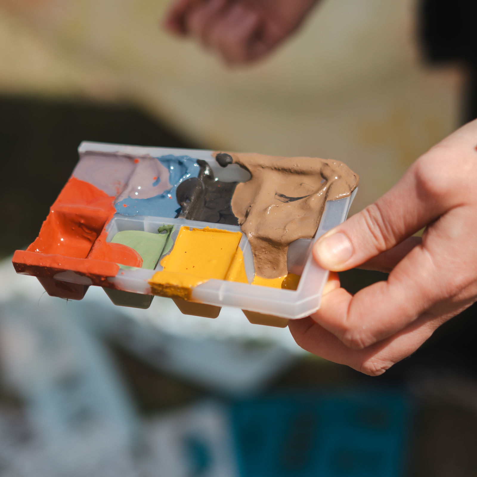 Hand mit einer Palette angerührter Lehmfarbe in Rot, Gelb, Grün, Blau und Braun