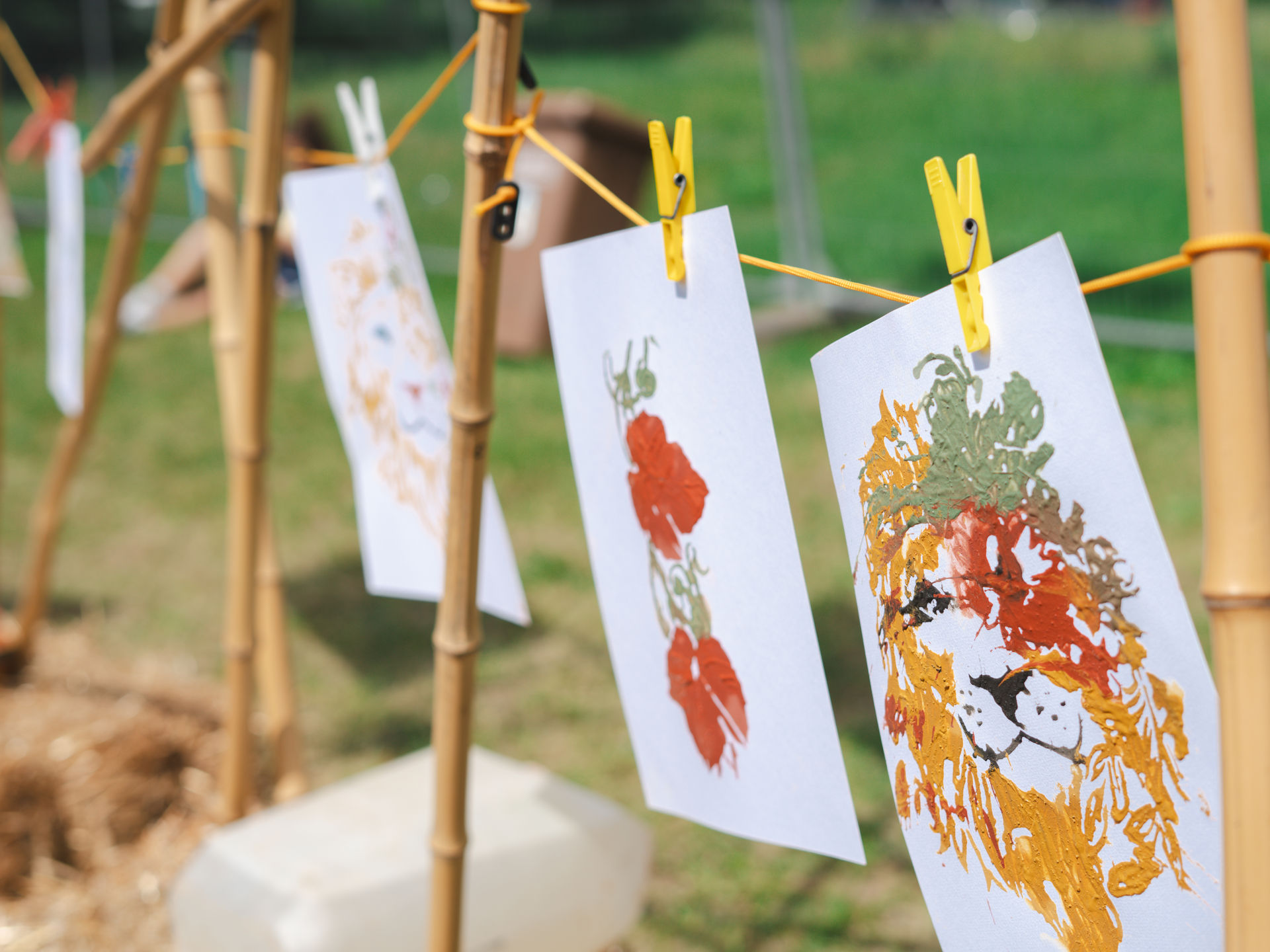 Mit Lehmfarben gemalte Kinderbilder – ein Löwe und Blumen – hängen mit Klammern an einer Leine im Freien
