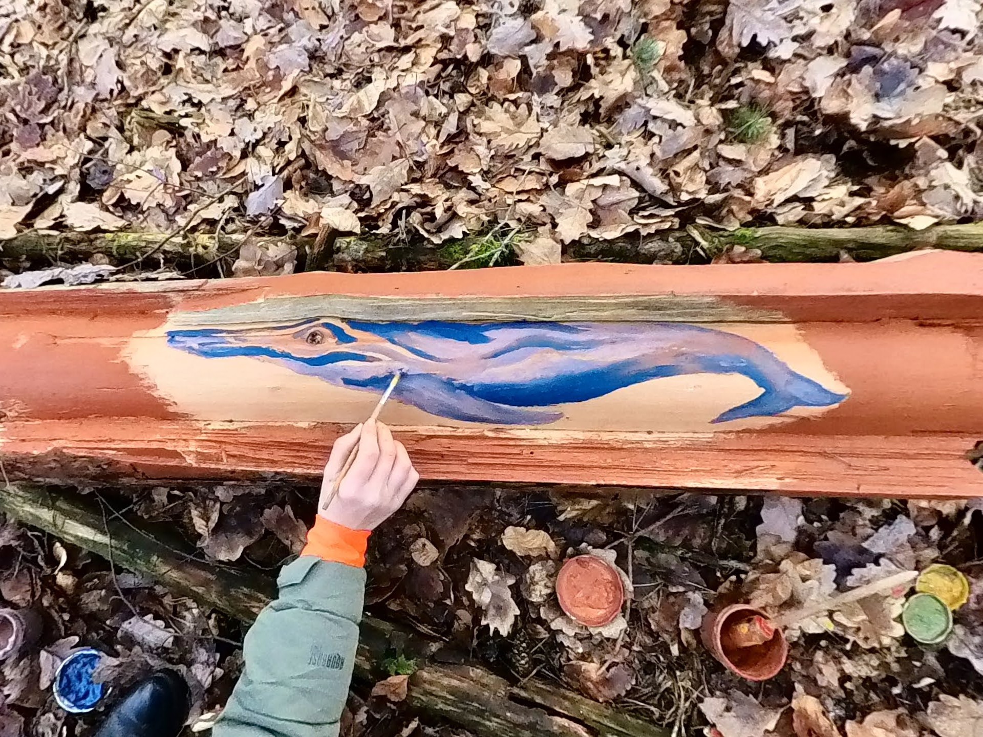Ein Kind malt mit dem Pinsel einen blauen Wal auf ein Stück Holz im Wald
