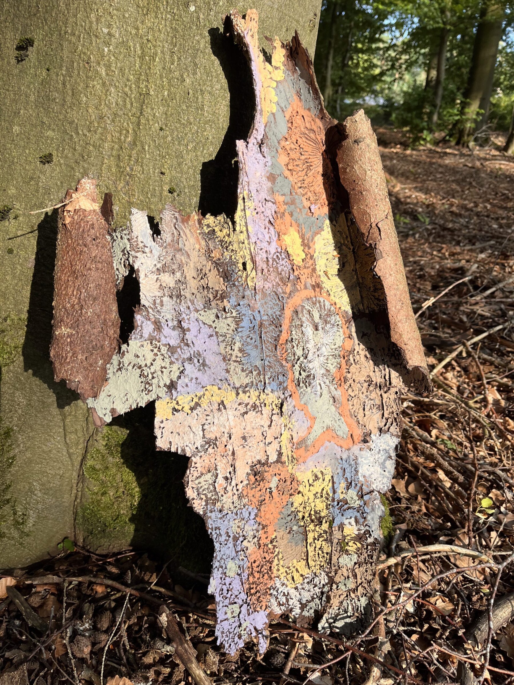 Ein Stück Baumrinde, bunt bemalt mit Mustern in Orange, Gelb und Flieder