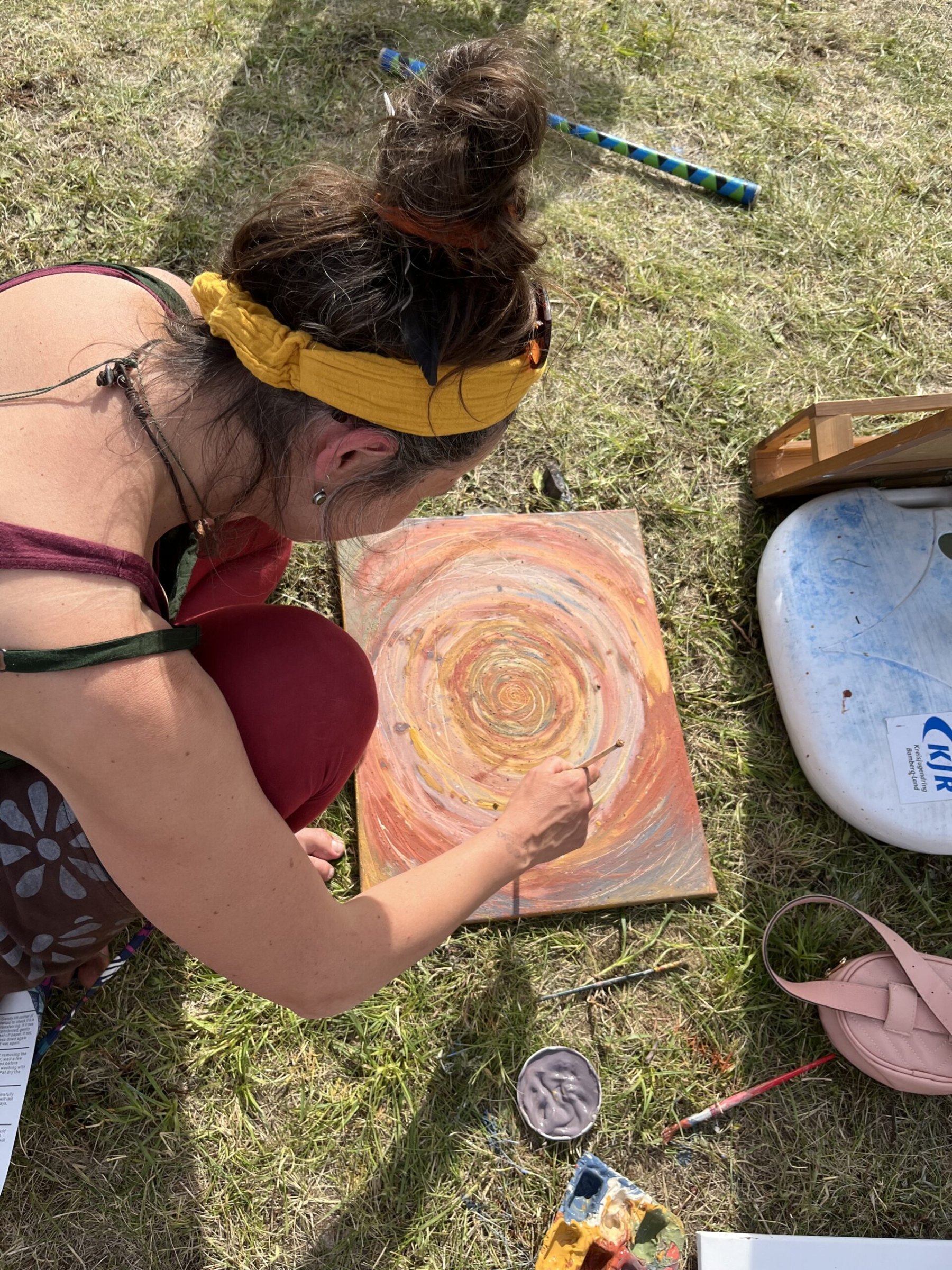 Eine Frau mit gelbem Haarband kniet im Gras und malt eine Spirale in Erdtönen auf eine Leinwand